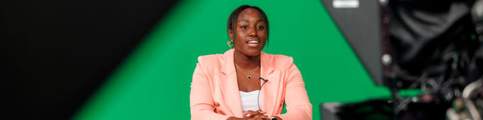 A Black woman in a pink blazer sits at a desk in a news studio with a green screen background.